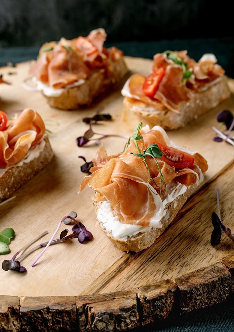 Plátky křupavého chleba s krémovým sýrem, tenkými plátky prosciutta, rozpůlenými cherry rajčátky a mikrozeleninou na dřevěném servírovacím prkénku.
