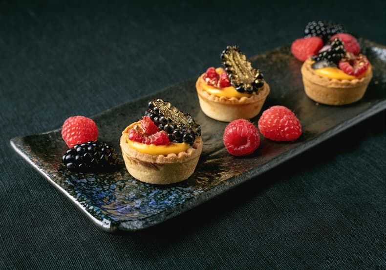 Tři malé tartaletky se žlutým krémem, zdobené ostružinami, malinami a zlatými lístky, podávané na černém obdélníkovém talíři s dalšími malinami a ostružinami.