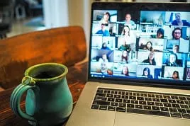 A close-up of a laptop displaying a virtual meeting with numerous participants on the screen for Google Ads training. A blue ceramic mug filled with coffee sits beside the laptop on a wooden table.