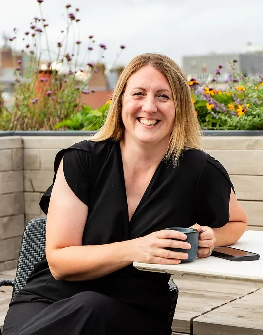 Charlotte Osborne sitting on a rooftop terrace, smiling while holding a blue mug. She is dressed in a black top and has shoulder-length blonde hair, surrounded by colorful flowers and an urban backdrop.