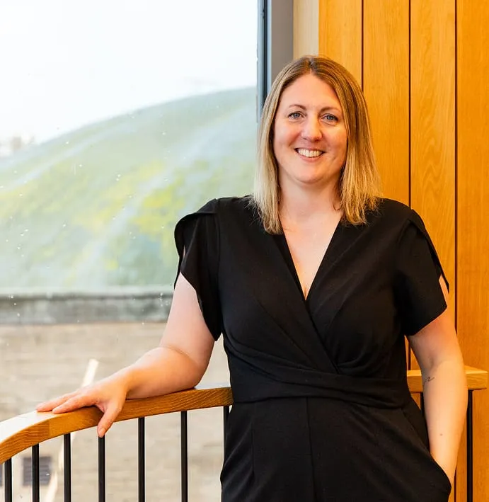 Charlotte Osborne wearing a black dress, standing beside a wooden railing in an office with a background of green hills visible through a window. She exudes a professional appearance.