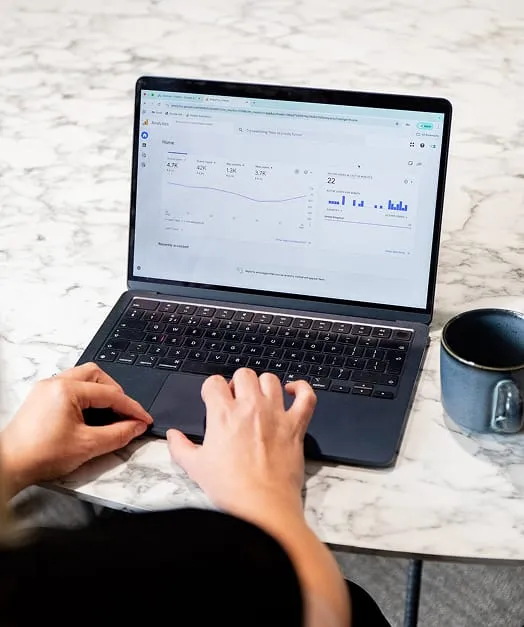 Charlotte Osborne using a laptop on a marble table, analysing Google Ads data displayed on the screen. A blue mug sits nearby, suggesting a casual workspace. The screen shows graphs and metrics related to PPC management.