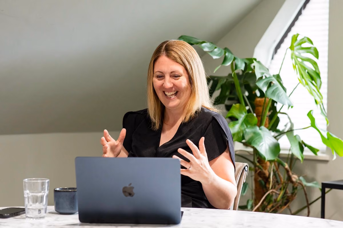 Charlotte Osborne smiling and gesturing while using laptop at a table with plants in the background.