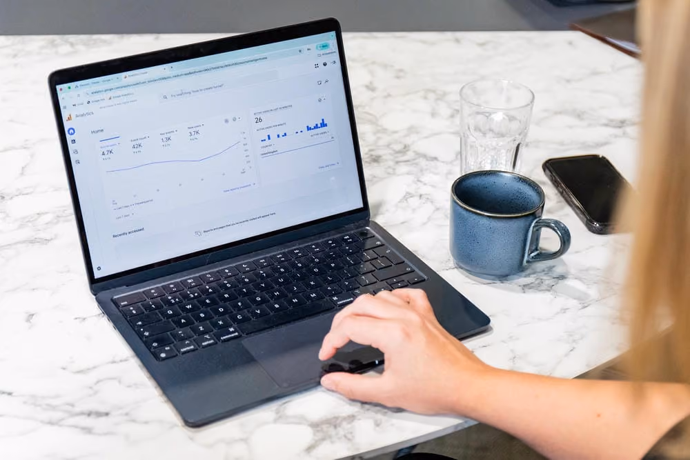 Person using a laptop showing web analytics data on a marble table with a cup, glass, and phone nearby.