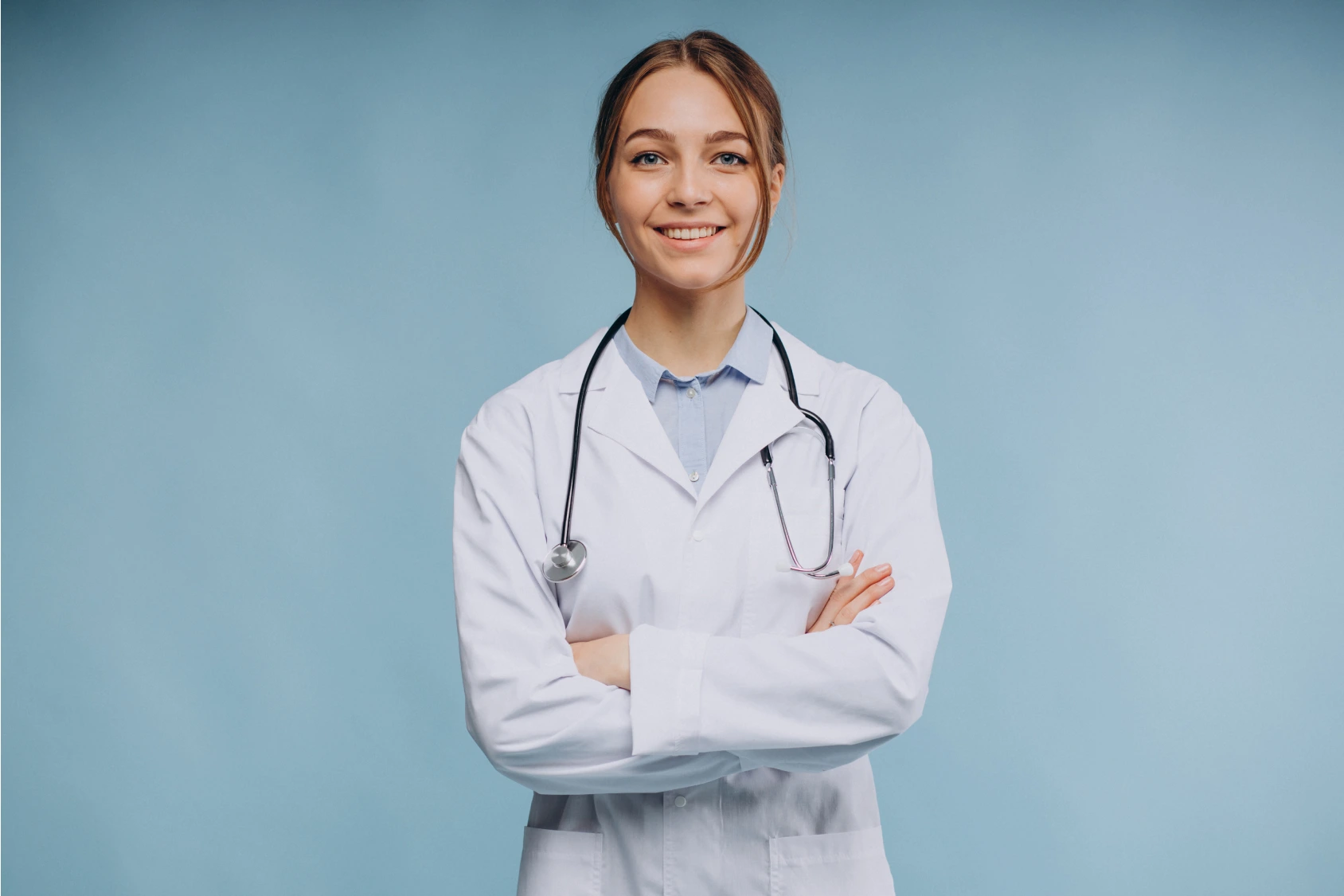 A female doctor smiling confidently, arms crossed, wearing a white coat with a stethoscope, standing in front of a blue background.