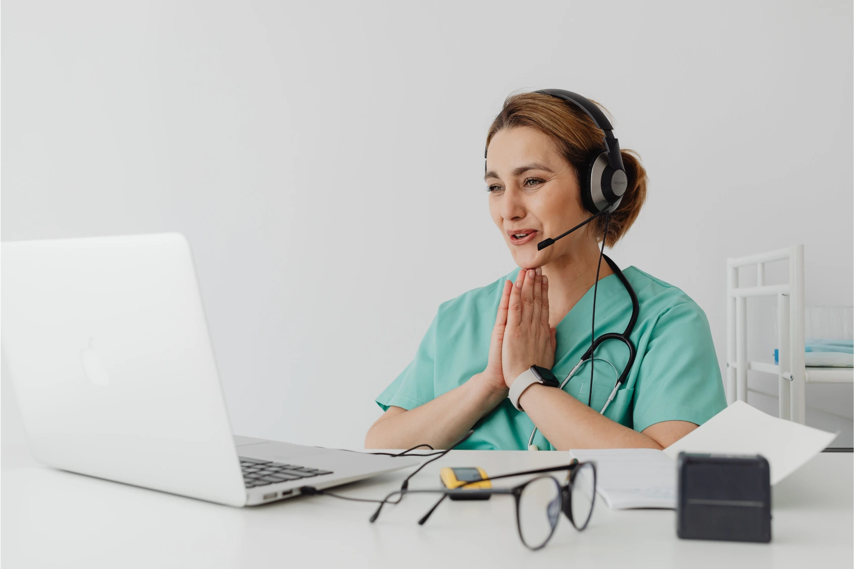 A female doctor wearing a headset and teal scrubs, conducting a telemedicine consultation on a laptop, with glasses and papers on the desk.