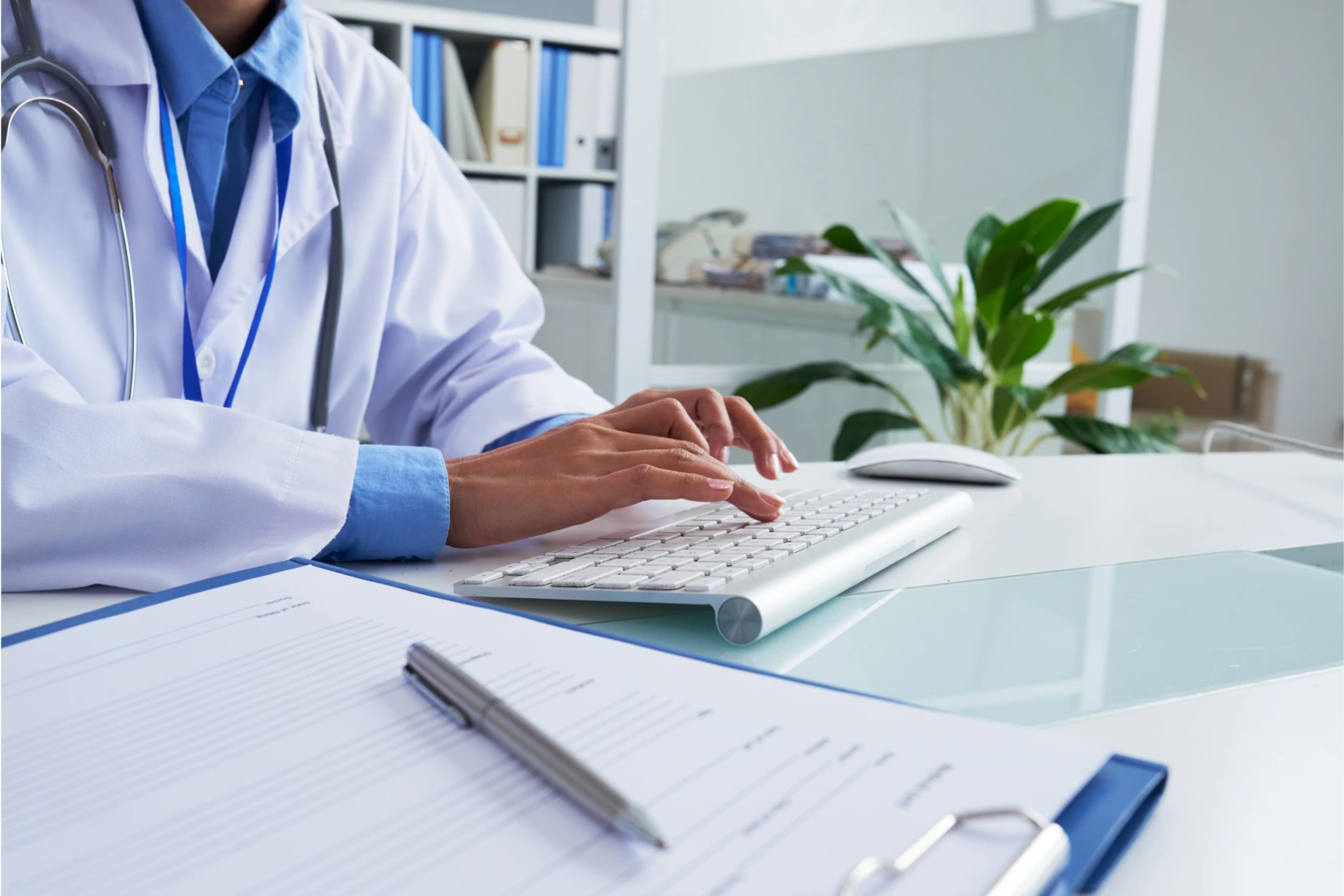 A doctor in a white coat typing on a keyboard in a bright, modern office, with medical documents and a pen on the desk in the foreground.