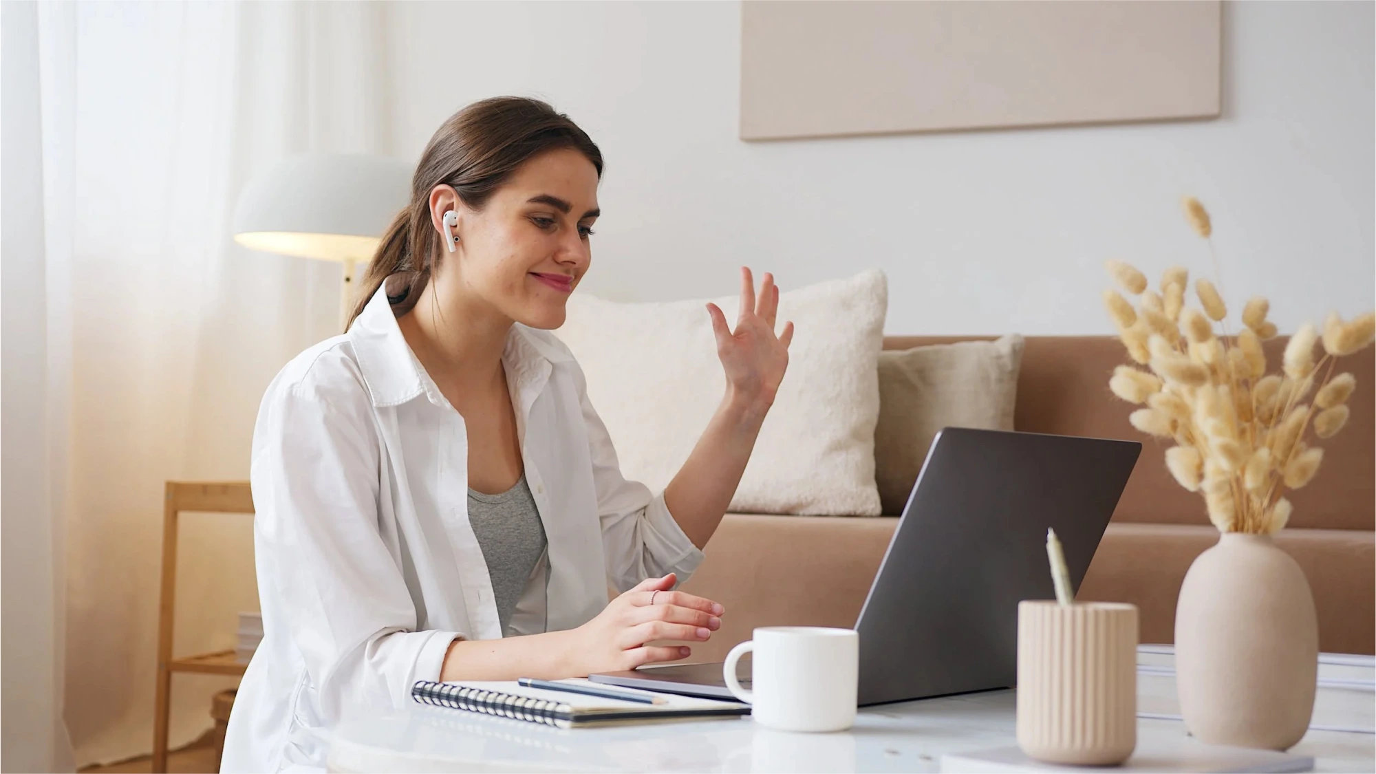 A woman conducting a virtual consultation on her laptop, smiling and waving while sitting at a table with a notebook and a cup nearby in a well-lit room.
