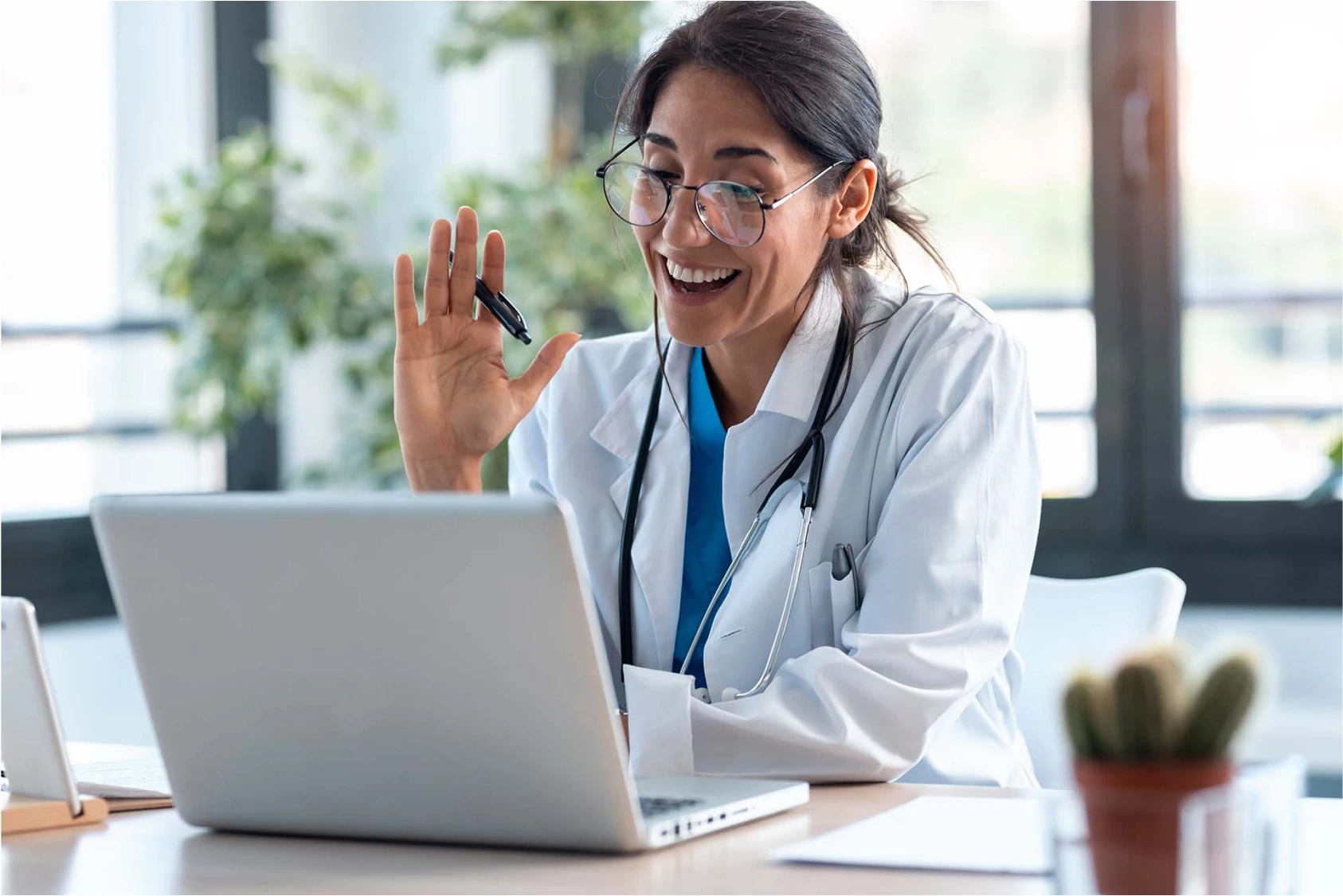 A female doctor sitting at her desk during a video consultation, smiling and waving at the screen while wearing a white coat and glasses.