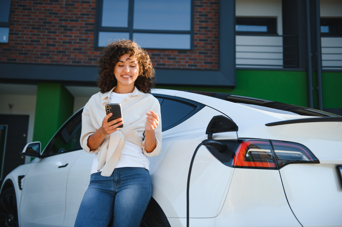 Junge Frau lehnt an einem Tesla Elektroauto und schaut auf ein Smartphone.