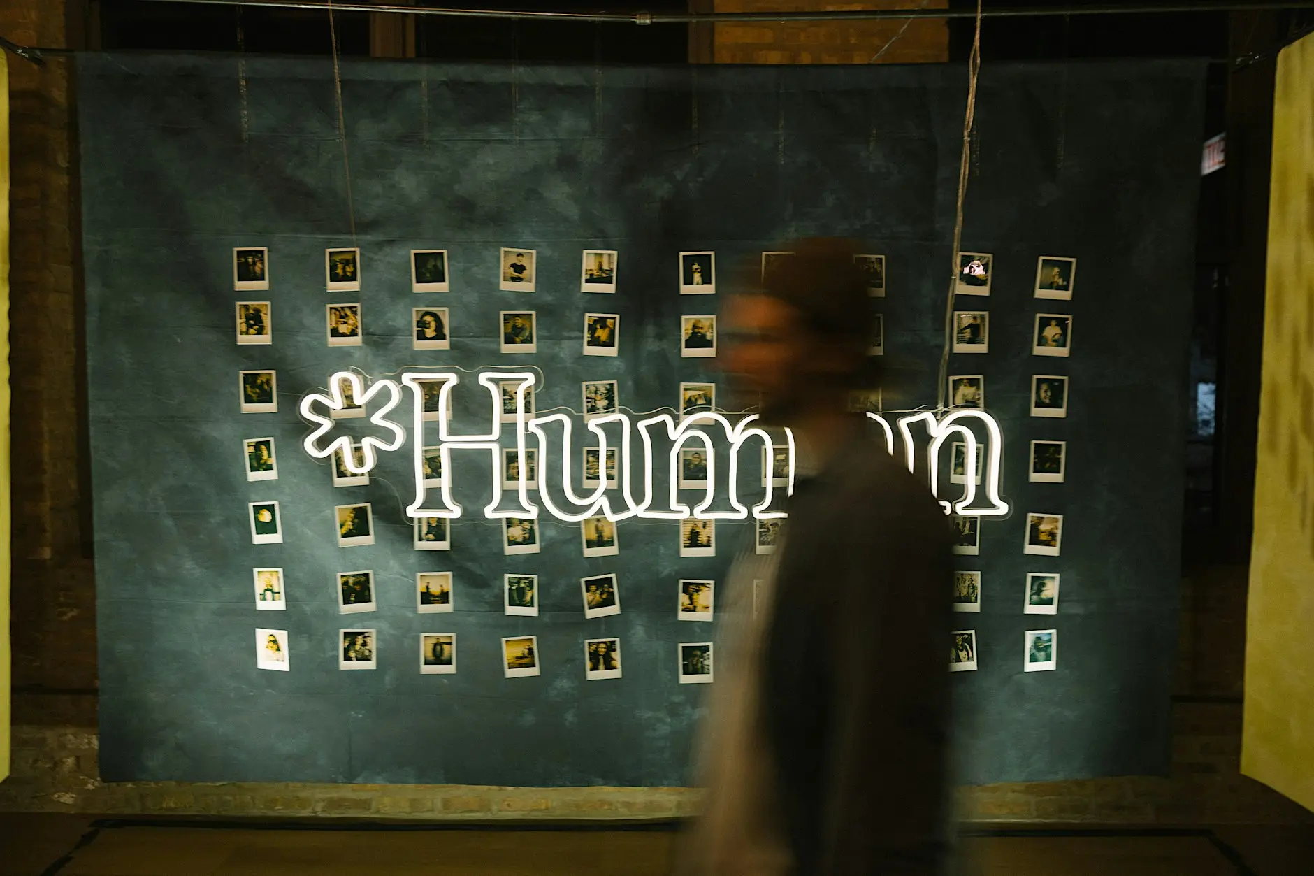 Blurred person walking past a lit *Human neon sign and a wall of instant photos at a *Human Chicago gathering