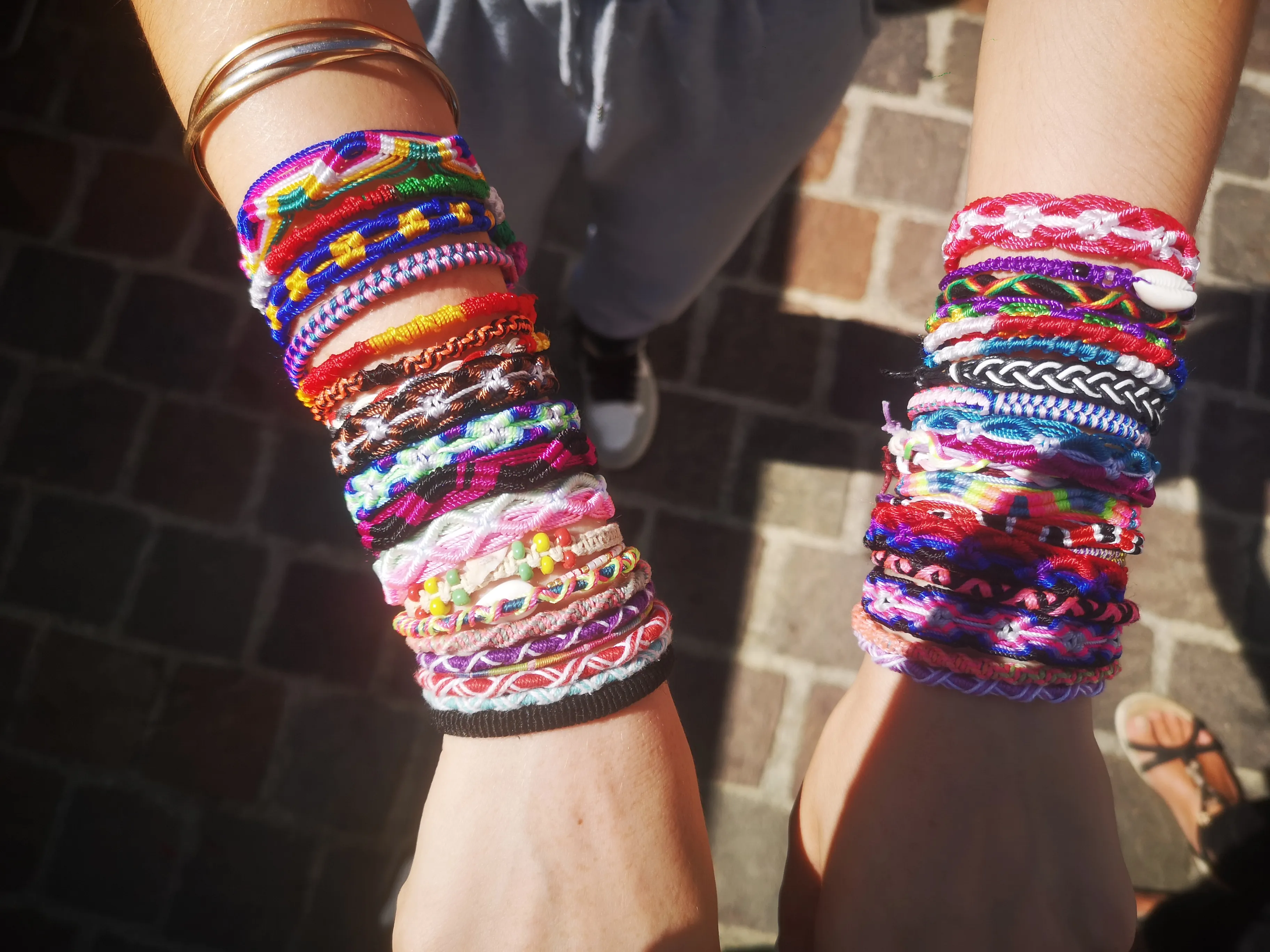 Poignets d'une personne portant de nombreux bracelets colorés en fils tressés et perlés, sur un fond pavé.