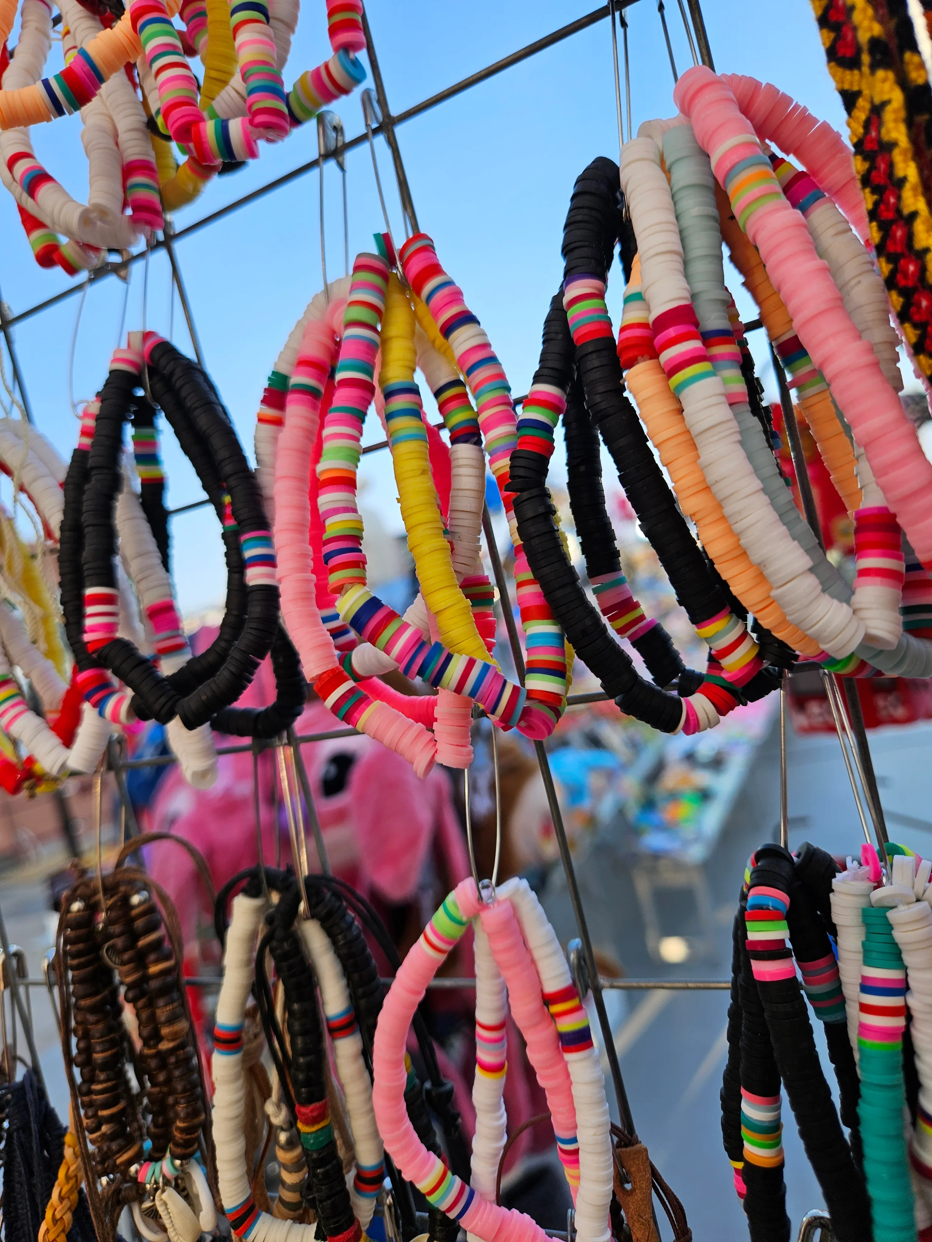 Bracelets colorés en perles plates suspendus sur un grillage métallique avec un ciel bleu en arrière-plan.