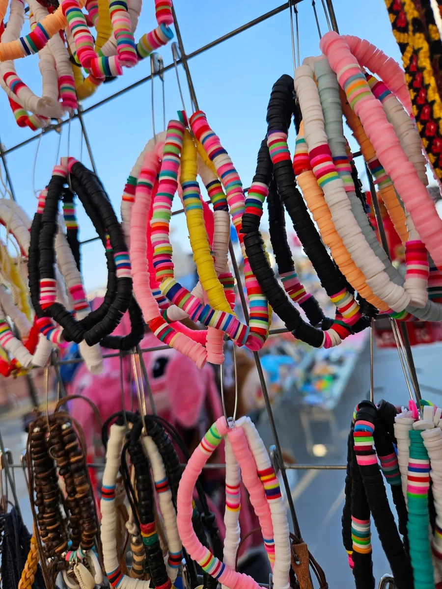 Bracelets colorés en perles plates suspendus sur un grillage métallique avec un ciel bleu en arrière-plan.