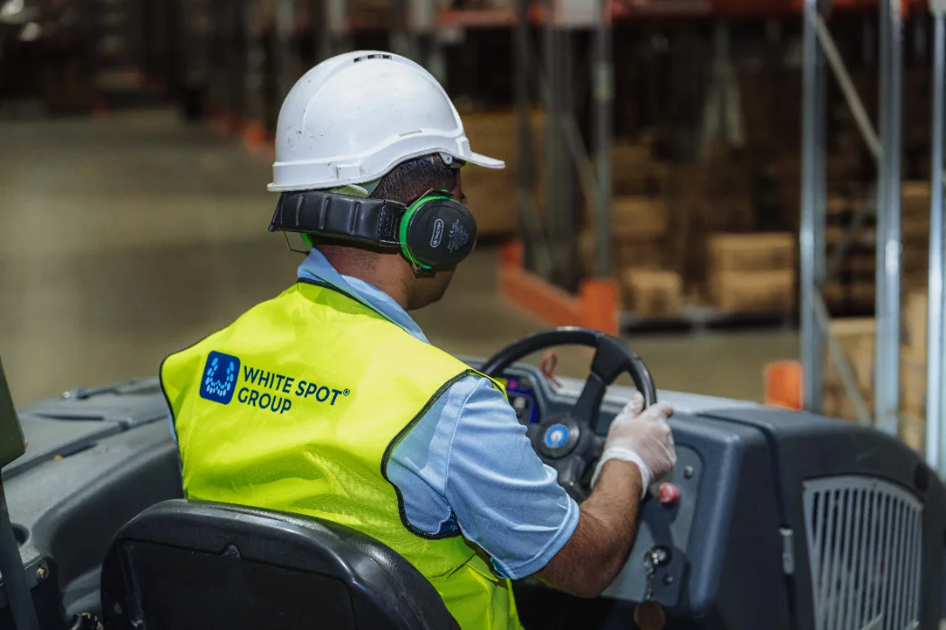 Warehouse worker wearing a white hard hat, green hearing protection, and a yellow safety vest with White Spot Group logo, driving a forklift inside a warehouse.