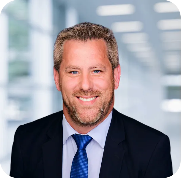 Smiling man with light brown hair and beard wearing a dark suit, light shirt, and blue tie against a blurred office background.