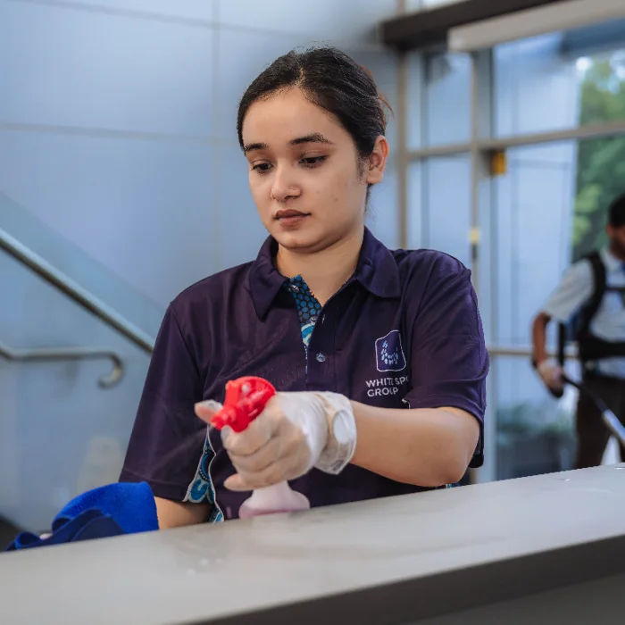 Woman in a dark blue uniform spraying cleaner on a surface while wearing gloves indoors.