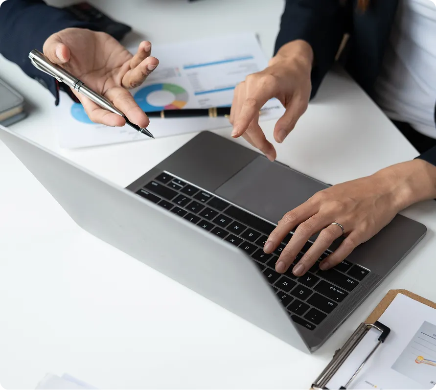 Hands of two people working on a laptop with business charts and documents on a white desk.