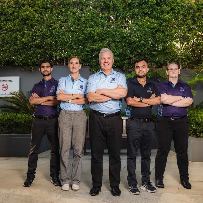 Three diverse factory workers in blue uniforms smiling with arms crossed inside an industrial warehouse.
