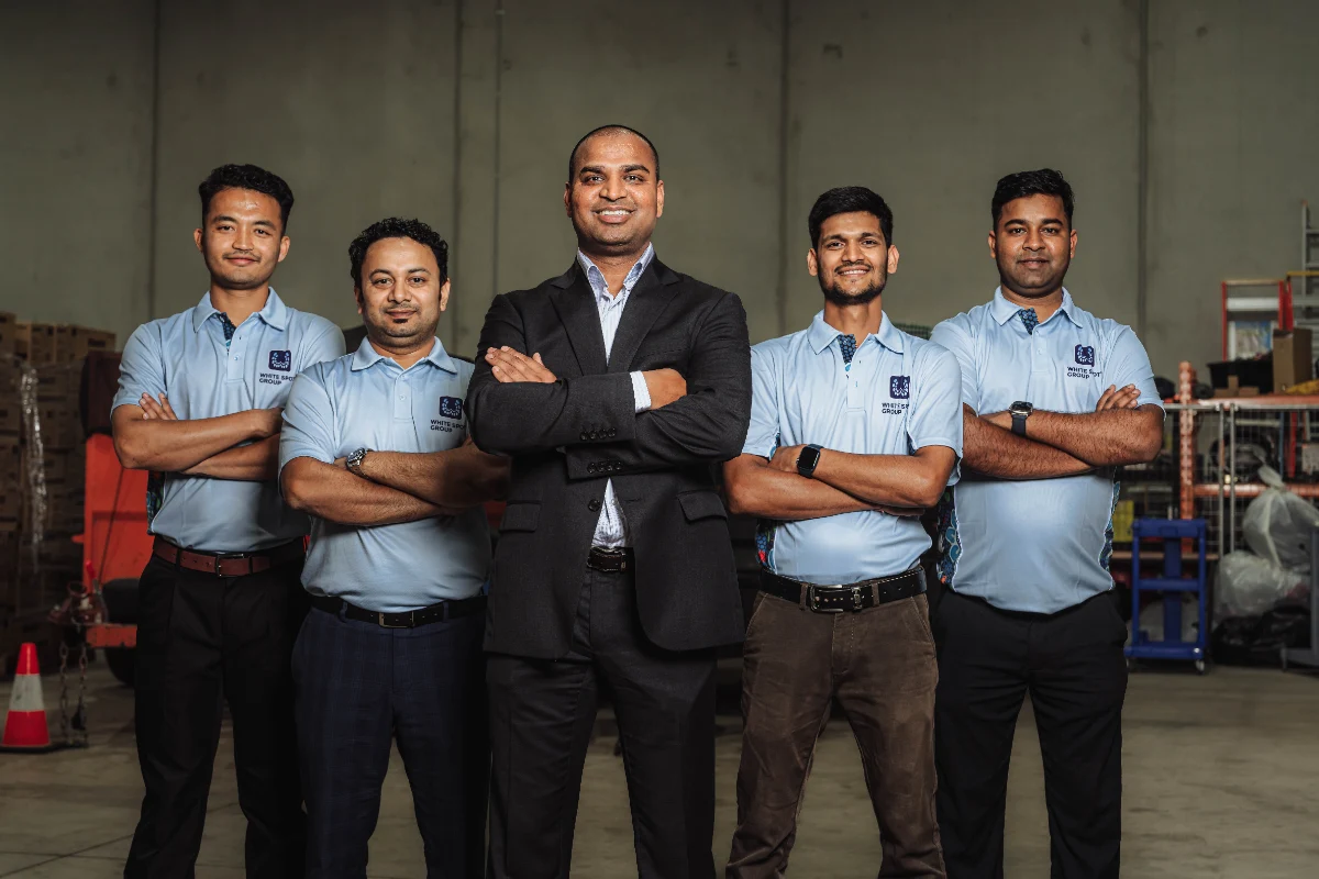 Five men standing with arms crossed in a warehouse, four in light blue polo shirts with 'White Spot Group' logos and one in a black suit.