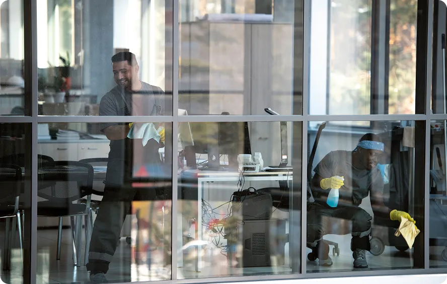 Two janitors cleaning the interior glass walls of an office with wiping cloths and spray bottles.