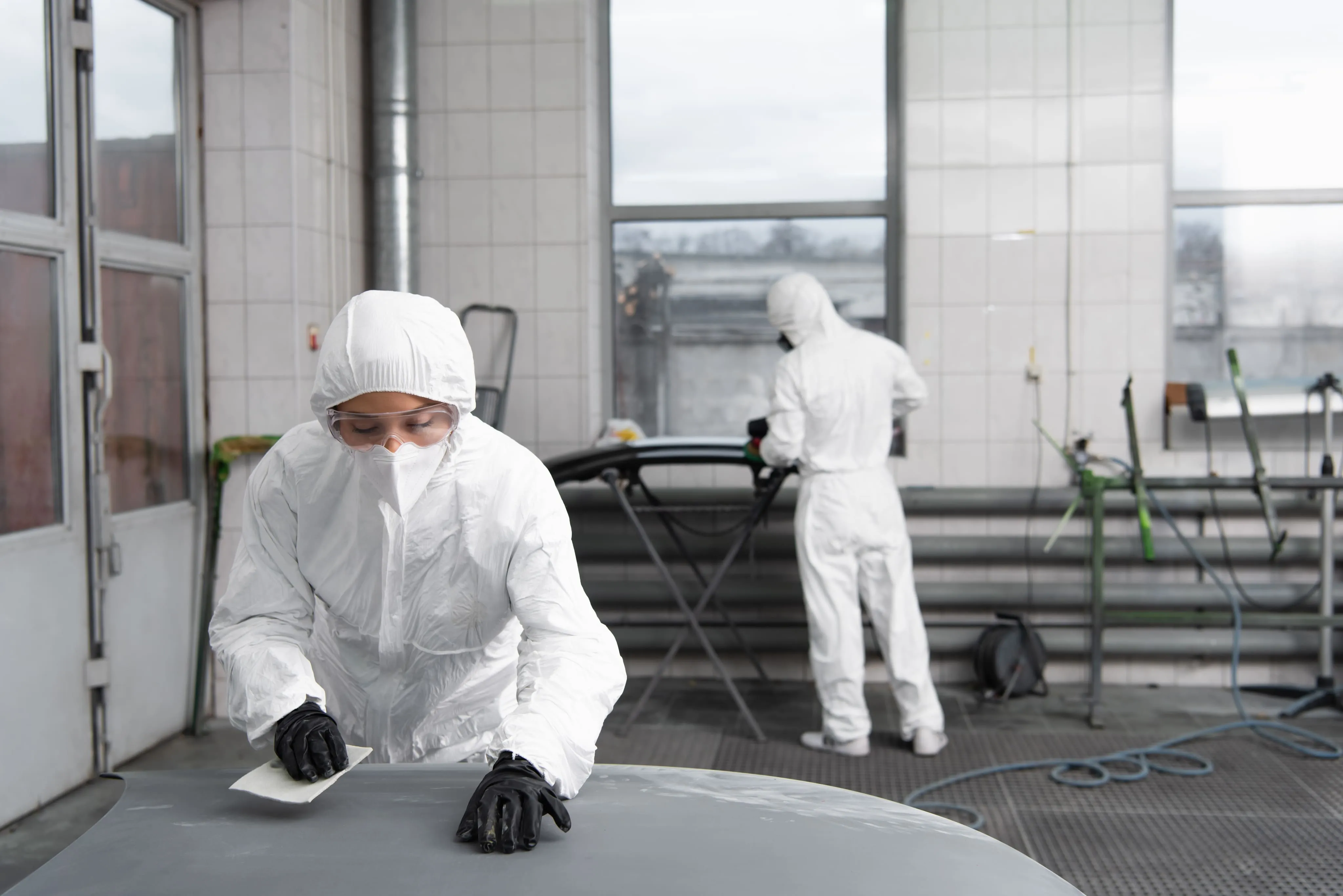 Two workers in white protective suits sanding and preparing car parts inside an auto repair workshop.