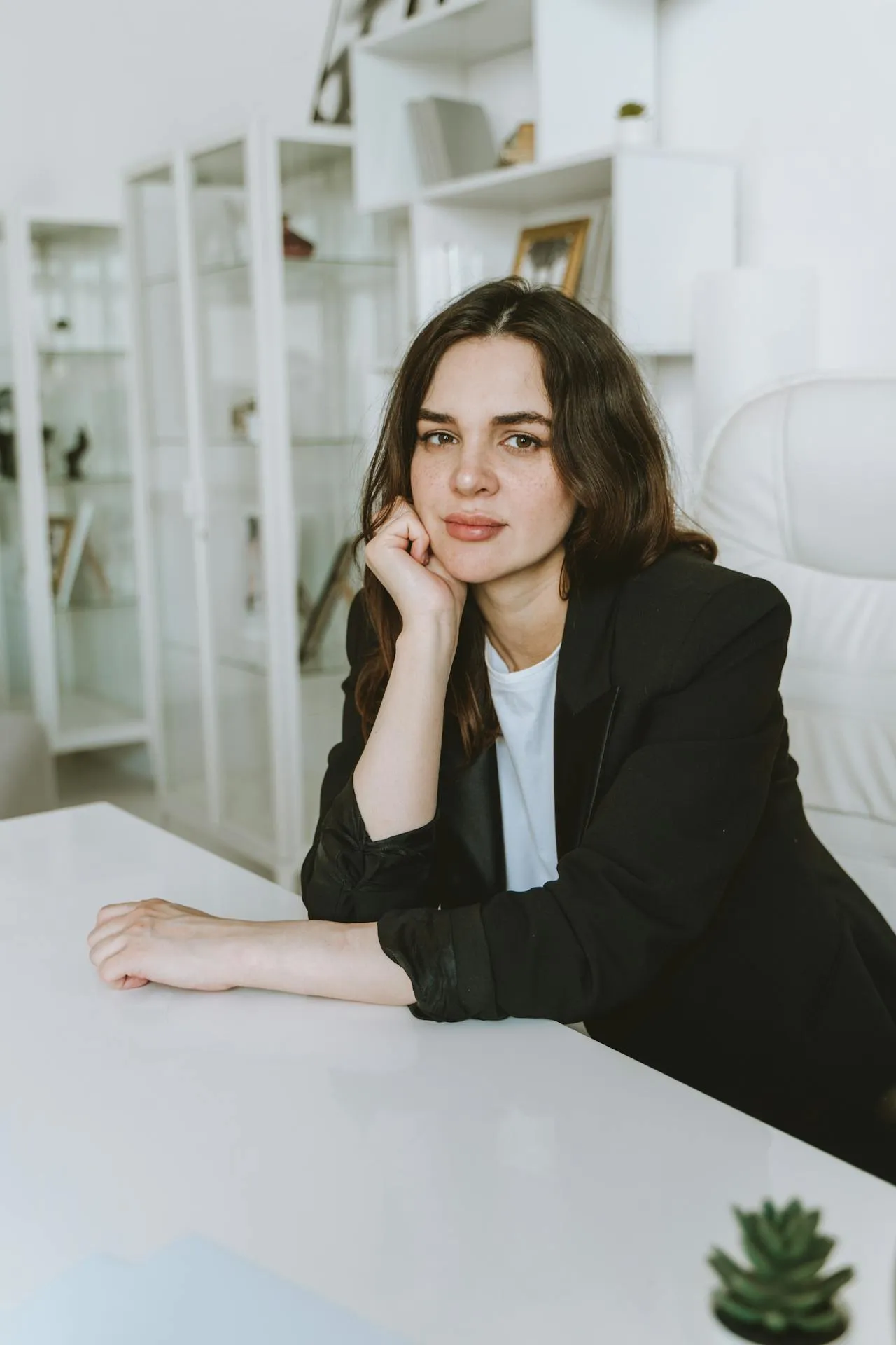 Woman with dark hair wearing a black blazer sitting at a white desk in a bright office.