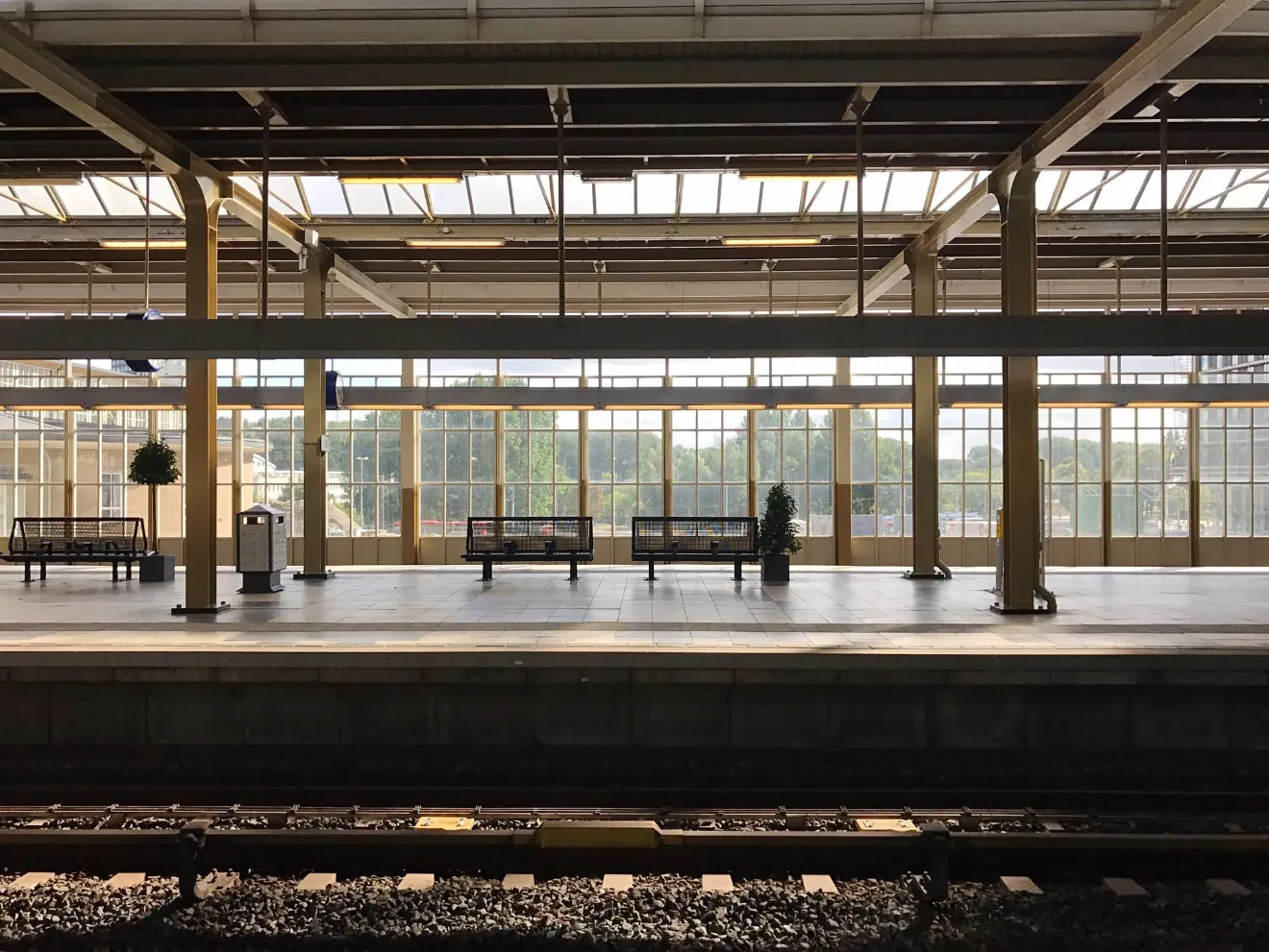 Empty train station platform with benches, plants, and large windows letting in natural light.