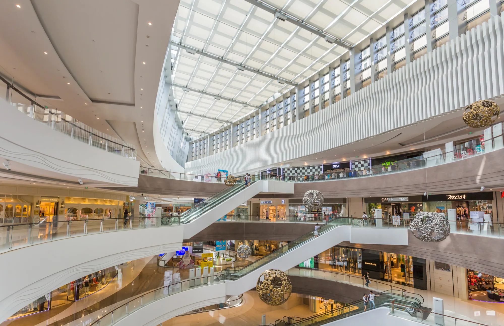 Interior view of a modern multi-level shopping mall with escalators, glass railings, hanging decorative spheres, and large skylights.