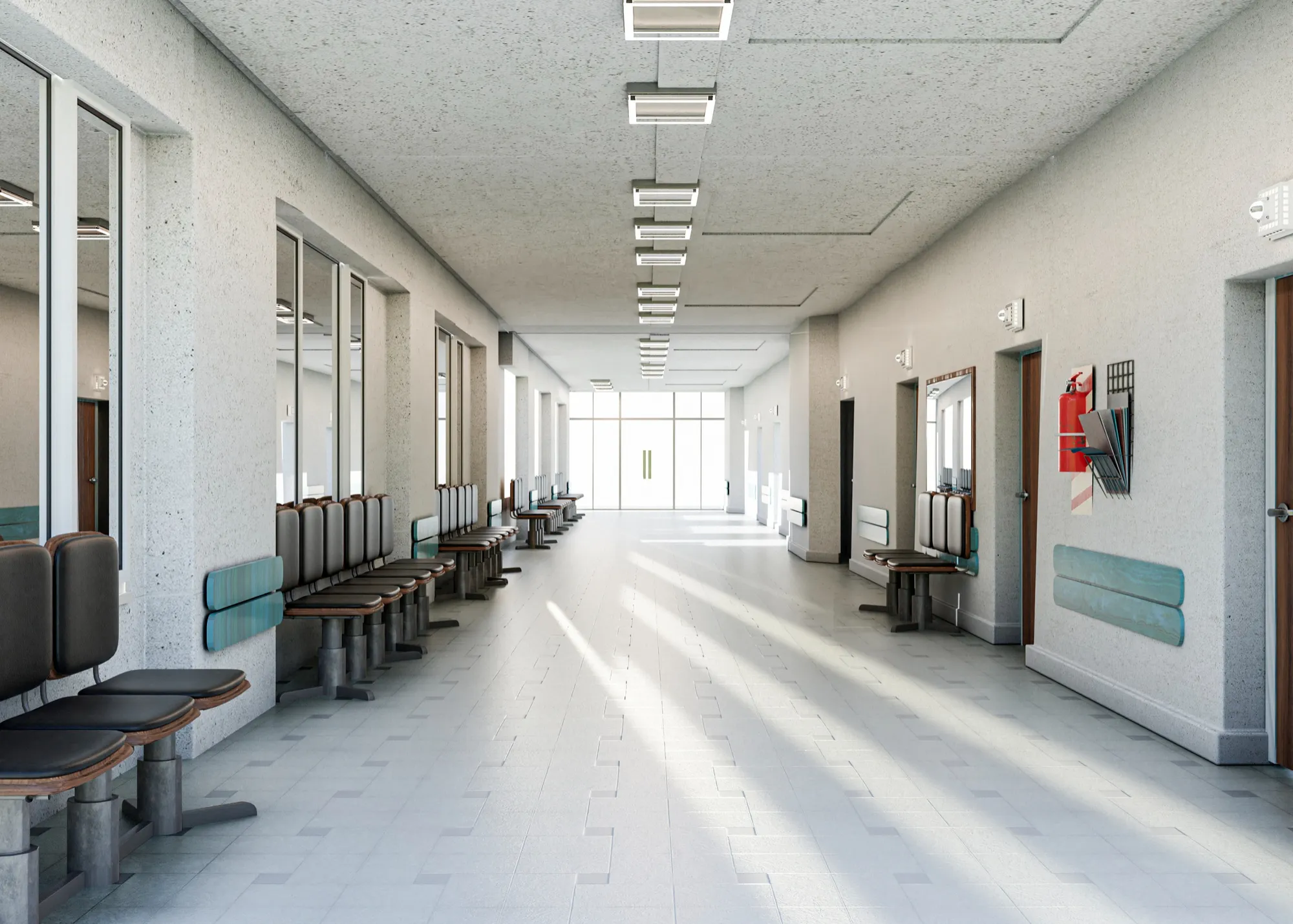 Bright empty hospital corridor with rows of chairs along both walls and large glass doors at the end.