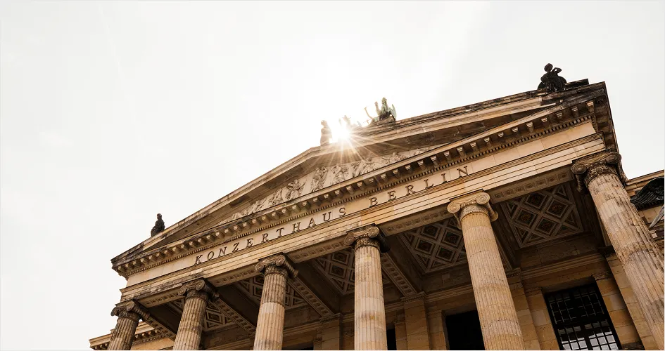 Low-angle view of the neoclassical Konzerthaus Berlin with the sun shining behind statues on the roof.