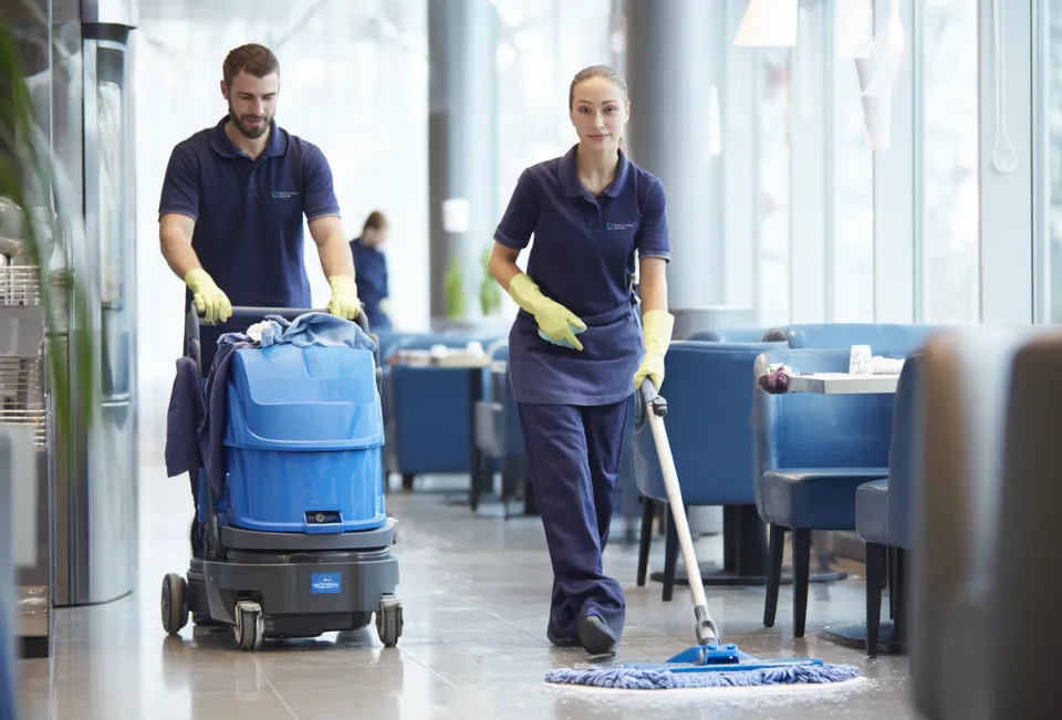 Two janitors in navy uniforms cleaning and mopping the floor in a modern restaurant with large windows and blue seating.