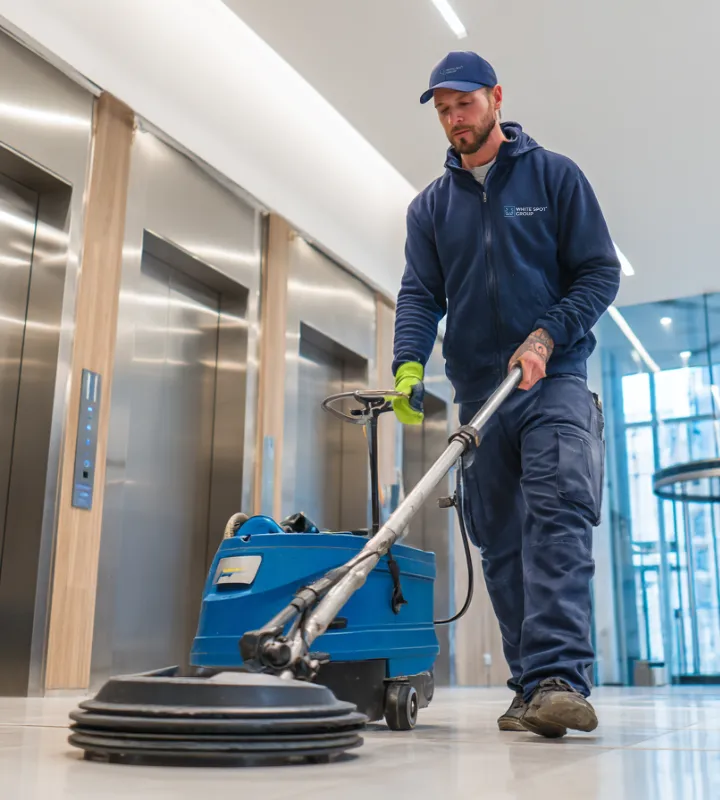Man in blue uniform using a floor cleaning machine in a modern elevator lobby.