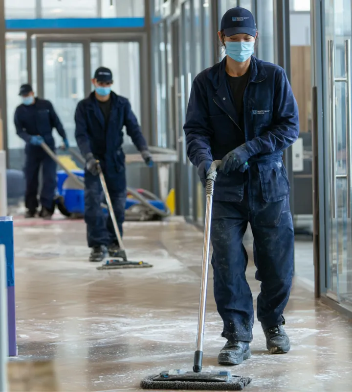 Three workers in masks and navy uniforms mopping a large indoor floor with cleaning equipment.