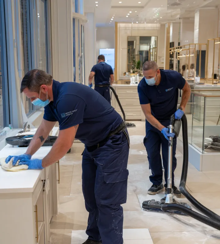 Three cleaning staff wearing blue uniforms, masks, and gloves, cleaning a bright, modern interior space; one wipes a countertop, another vacuums the floor, and a third works in the background.