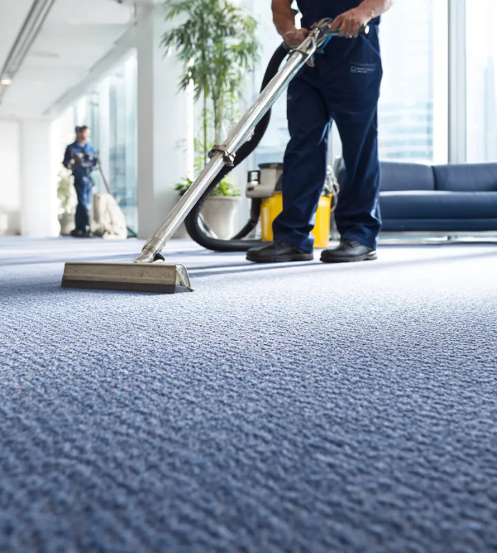 Two professional cleaners in blue uniforms cleaning a large office carpet with industrial carpet cleaning machines.