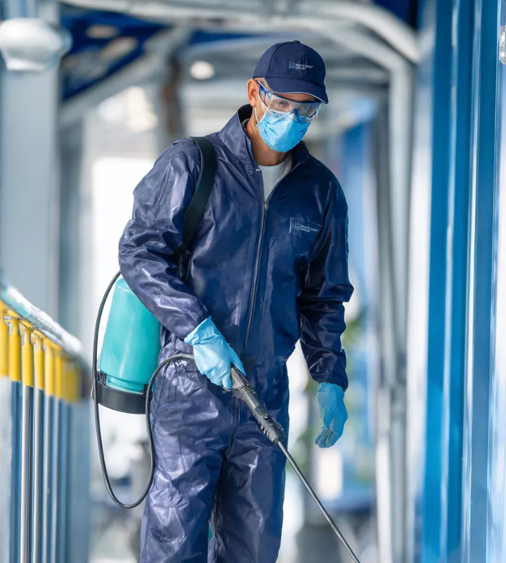 Person wearing full protective gear with mask, gloves, goggles, and a blue suit disinfecting an area using a sprayer.