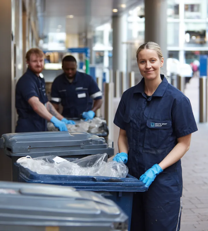 Three sanitation workers in dark blue uniforms and gloves handling large outdoor waste bins on a city street.