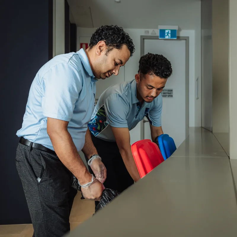 Two men in light blue shirts cleaning and handling waste bins indoors.