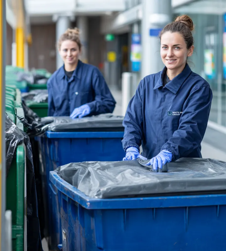 Two women in blue uniforms and gloves handling large blue covered trash bins outdoors.