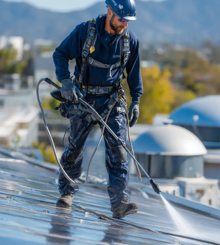 Worker wearing safety harness and helmet pressure washing a rooftop under clear sky.