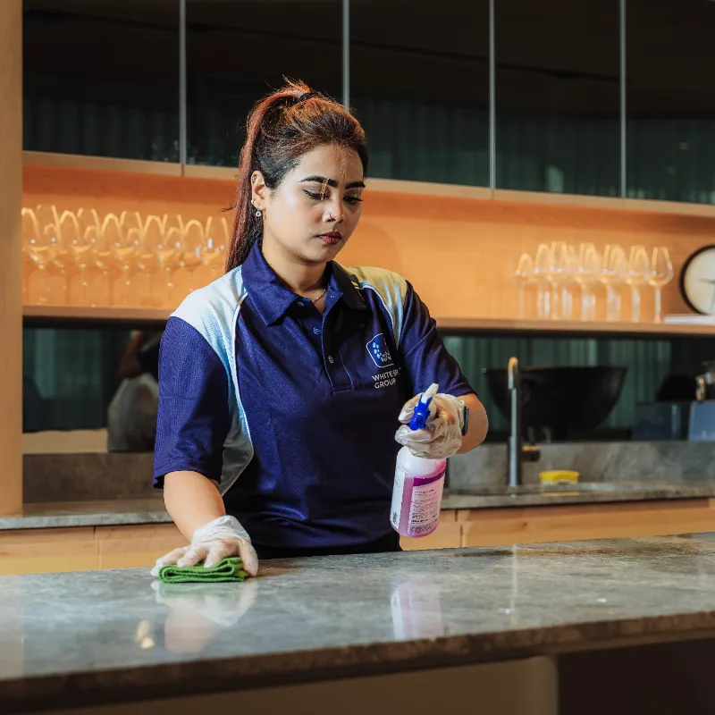Woman wearing gloves cleaning a countertop with a cloth and spray bottle in a modern kitchen or bar area.