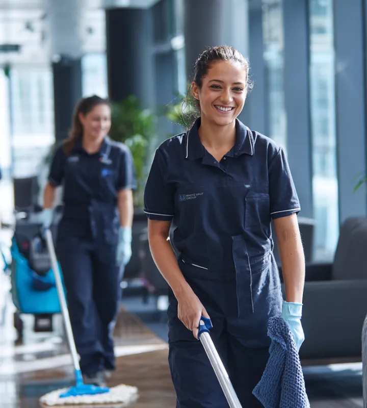 Two smiling female janitors in navy uniforms cleaning a modern office space with mops.