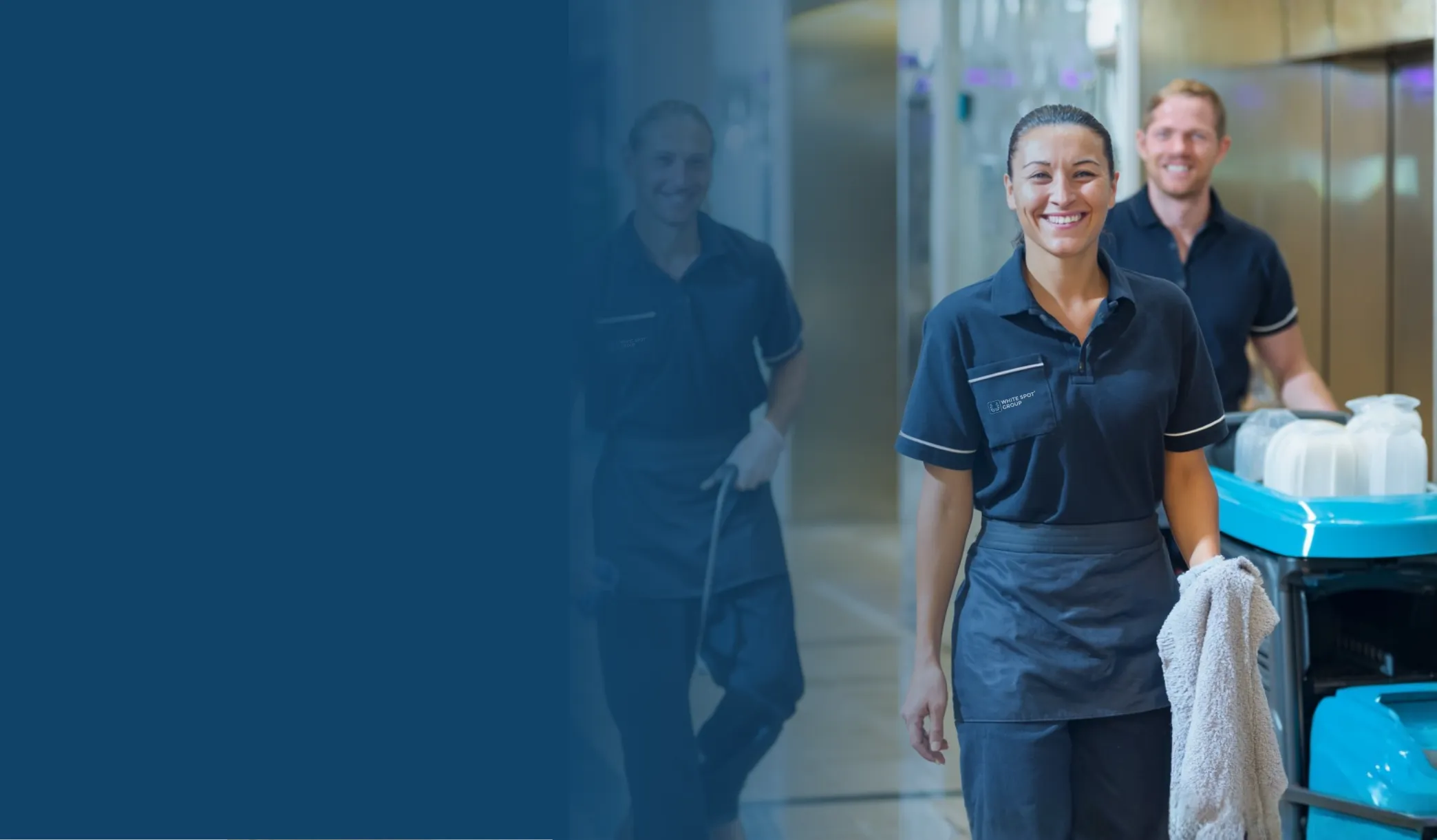 Smiling cleaning staff in navy uniforms with cleaning cart and cloth in a hallway.