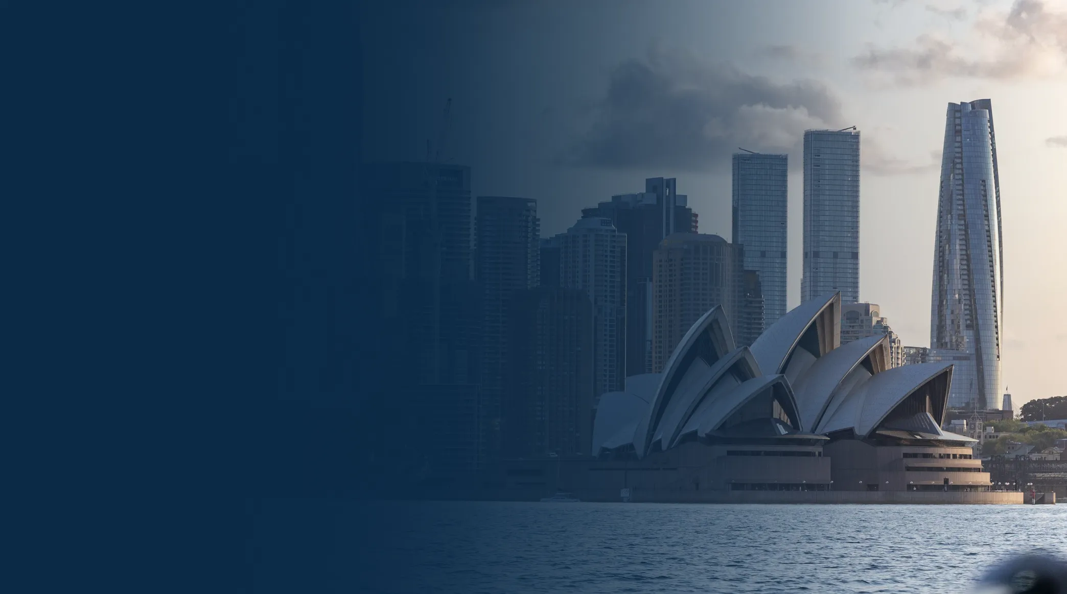 Sydney Opera House with city skyscrapers in the background by the water at dusk.
