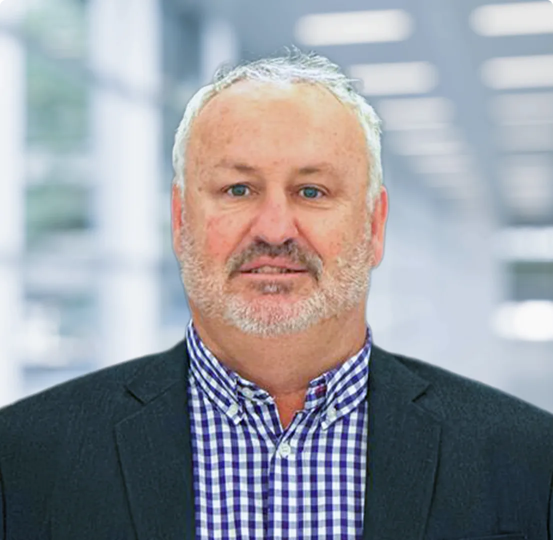 Middle-aged man with short gray hair and beard wearing a dark blazer and blue checkered shirt against a blurred office background.