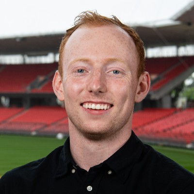 Smiling young man with red hair in a black shirt standing in a sports stadium.