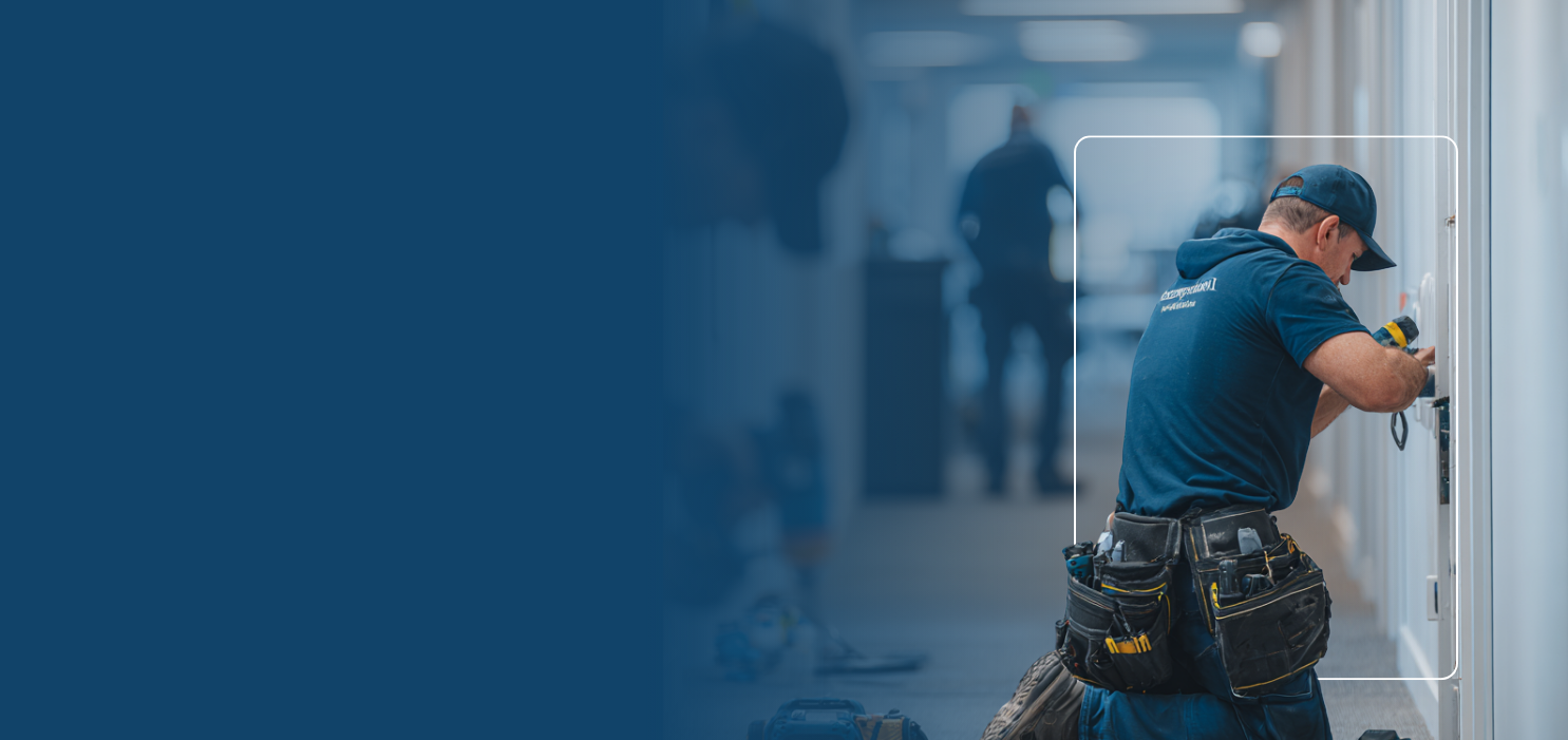 Worker wearing a tool belt and cap using a power drill on a white wall in a hallway.