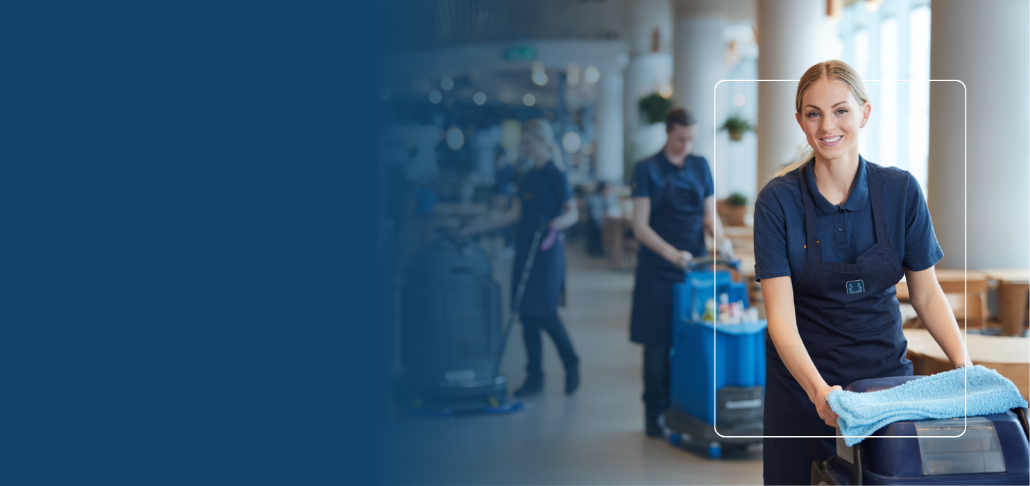 Smiling woman in navy cleaning uniform pushing a janitorial cart with cloth, with two other cleaning staff working in the background.