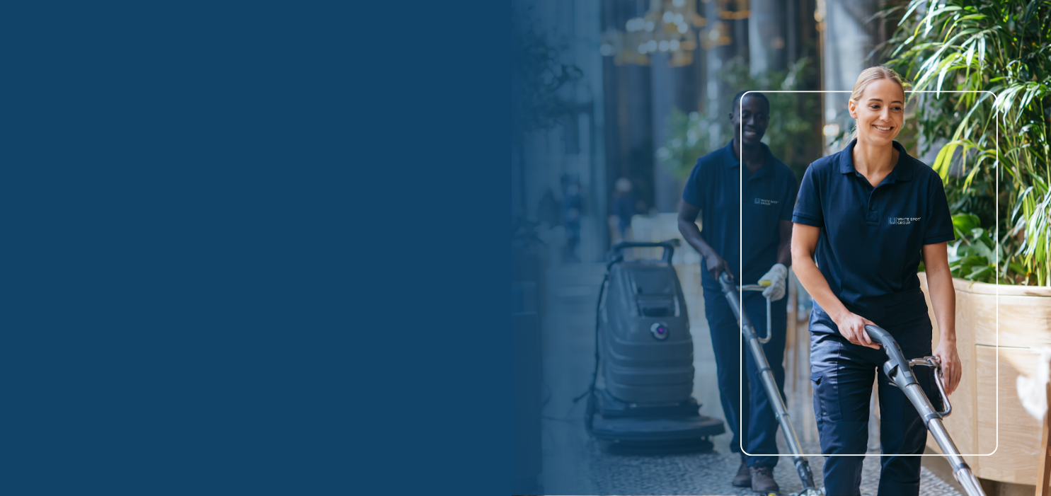 Two professional cleaners in navy uniforms operating floor cleaning machines in a bright indoor space with plants.